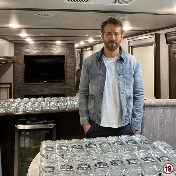 Ryan Reynolds in a blue jacket standing indoors with bottles of Aviation Gin, related to net worth and business decisions.