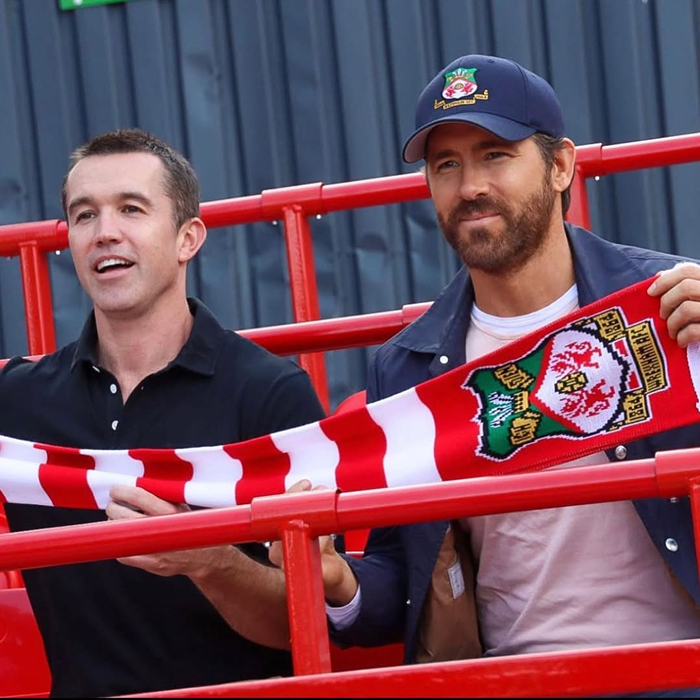 Two men holding a red and white scarf at a sports event, one wearing a blue cap Ryan Reynolds. Two men holding a red and white scarf at a sports event, one wearing a blue cap Ryan Reynolds.