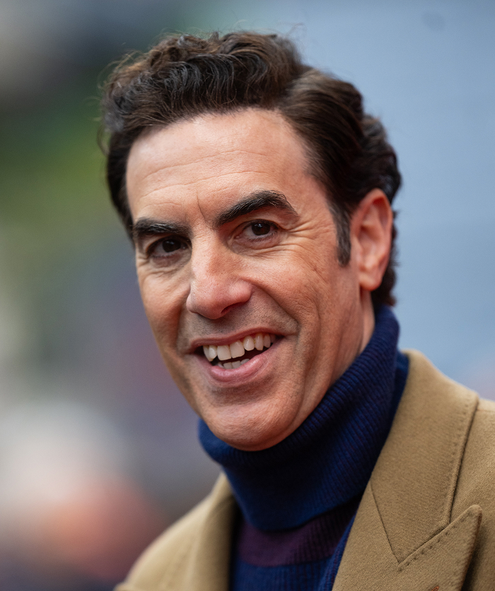 A celebrity smiling in a tan coat and blue turtleneck, epitomizing attractiveness beyond the screen.