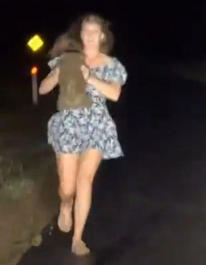 A woman in a floral dress holding a baby wombat at night on a road.