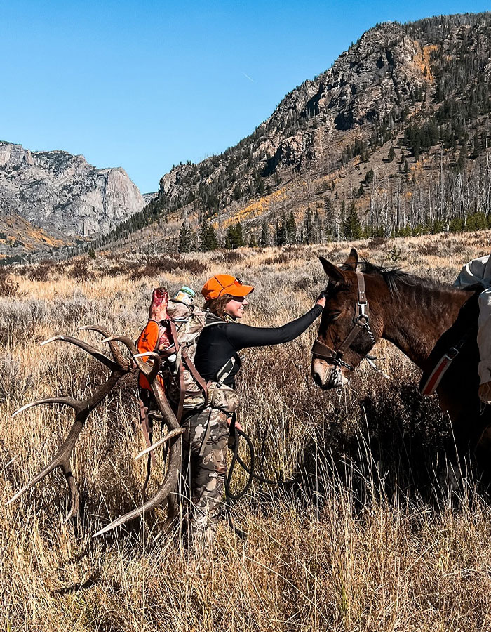 Woman in camo gear with antlers on her back petting a horse in a mountainous landscape.