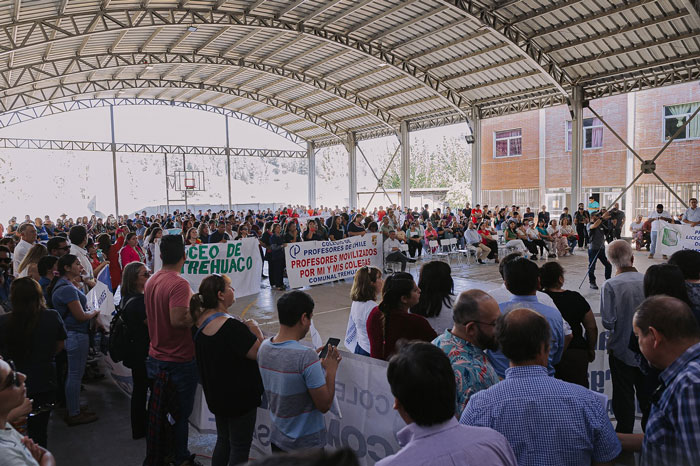 Large group gathered in a covered area, holding signs in support of a teacher after an incident involving a student.