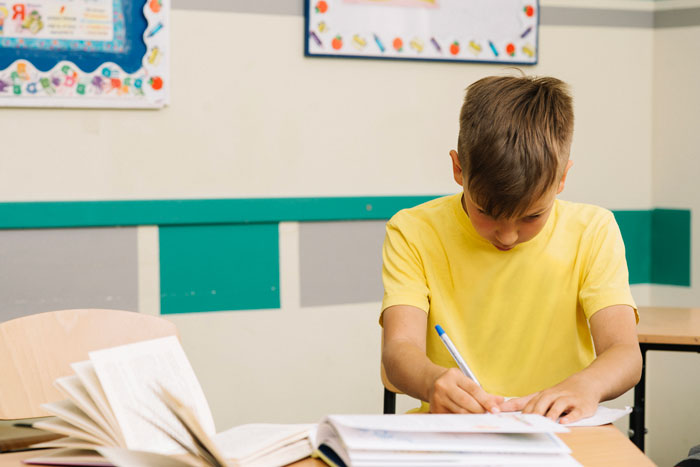 A teenage boy in a yellow shirt writes in a classroom, focusing on his work, surrounded by open books.