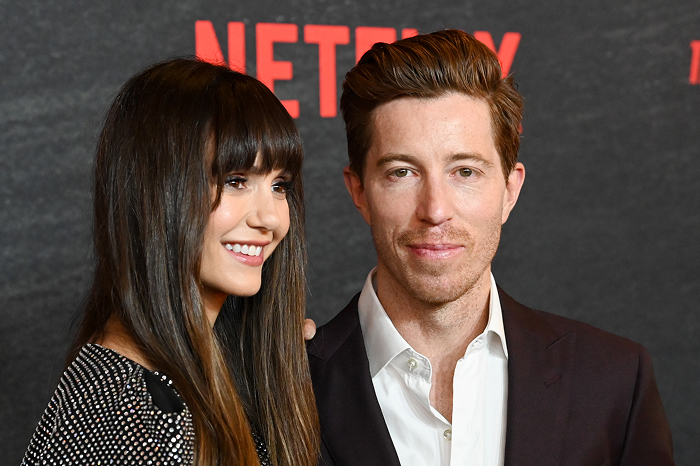 Shaun White and Nina Dobrev smiling at a Netflix event, she in a patterned dress and he in a dark suit.