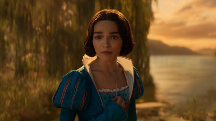 Actress in blue dress by a lake at sunset; Disney's Snow White scene.