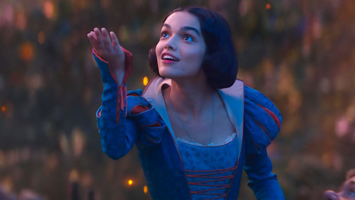 Actress in a blue dress as Snow White, smiling and reaching out against a forest backdrop, representing Disney's Snow White.