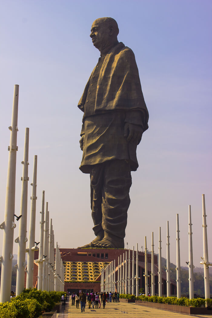 Iconic statue of a historical figure standing tall against a clear sky, surrounded by visitors and columns.