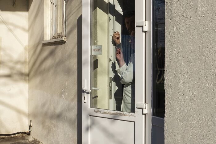 A man in white coat comically stuck behind a glass door, casting a playful look outside on a sunny street.