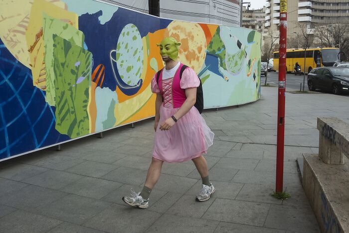 Person in a Shrek mask and pink dress walking by colorful street art, capturing a comedic street photo moment.