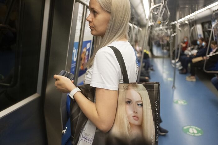 A woman on a subway with a tote bag featuring a face that humorously aligns with hers, creating a comic street photo moment.