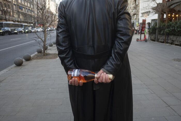 Street photo of a person in a black coat holding a bottle behind their back, capturing a humorous moment.