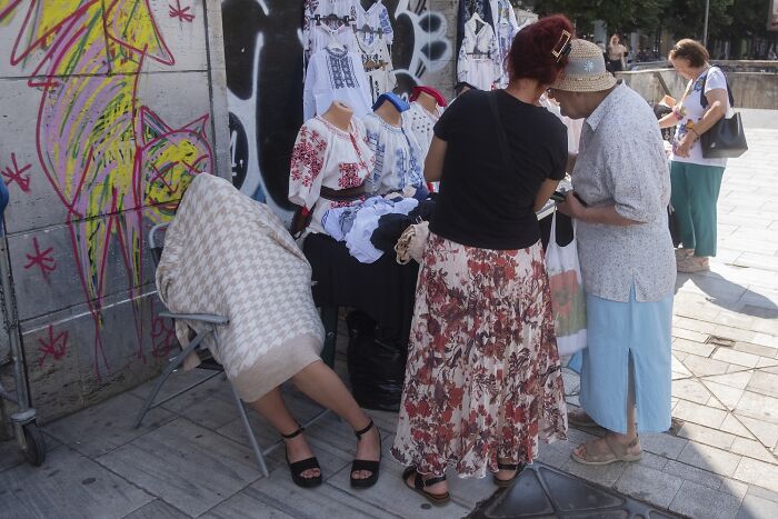 People browsing a street vendor's clothing stand, with a humorous twist as a hidden person behind a blanket adds comedy.