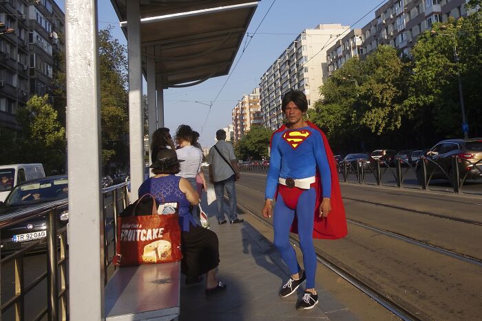 Man in Superman costume at a busy street tram stop, capturing a perfectly timed comedic street photo.