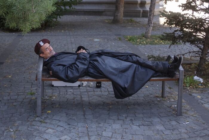Man in leather coat and beret humorously naps on a park bench, showcasing perfectly timed street photography comedy.