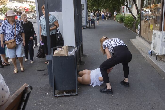 A woman in striped shirt helps another woman on the sidewalk, creating a comedic street photography moment.