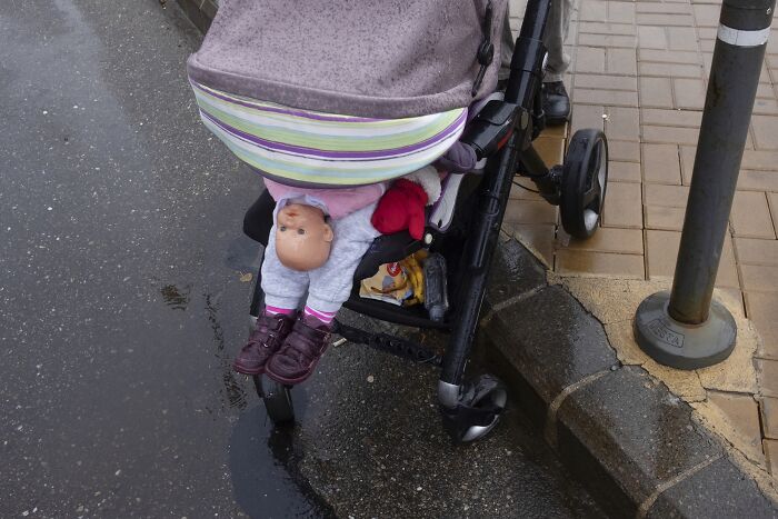 A doll humorously placed under a stroller by the street, creating a comedic moment in everyday street photography.
