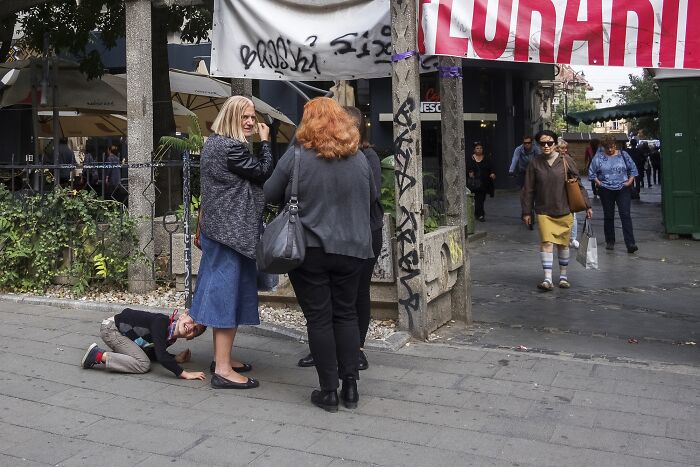 "Perfectly timed street photo capturing people chatting while a child playfully crouches on the sidewalk."