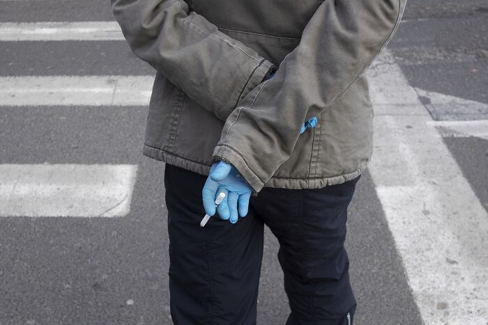 Person in a street holding a cigarette behind their back, wearing blue gloves and a green jacket, capturing a comedic moment.