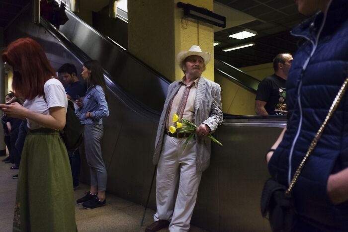 Elderly man in a hat holding flowers, leaning by an escalator in a busy street photo.