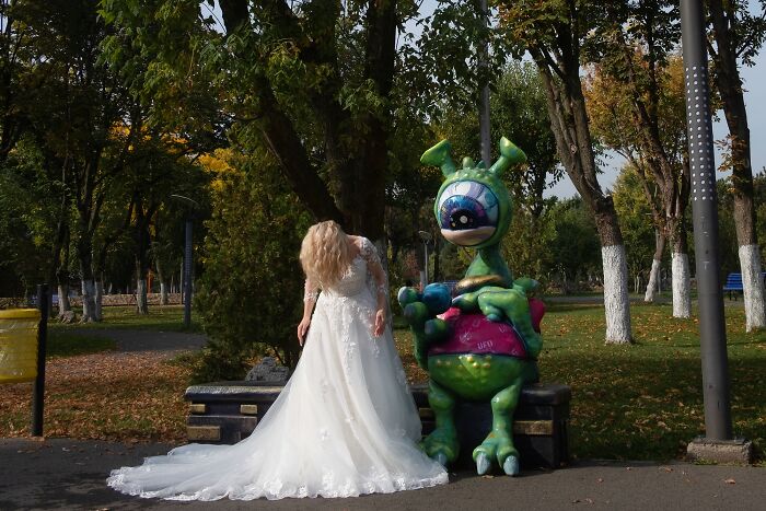 Bride in white dress bending over next to a colorful alien statue in a park, capturing a perfectly timed street photo moment.