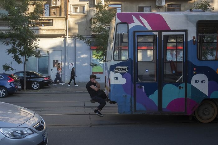 Man hangs onto a colorful tram on a street, capturing a perfectly timed comedic moment.