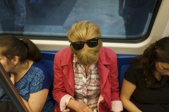 Street photo of a subway passenger with sunglasses on their hair resembling a face, creating a comedic everyday scene.