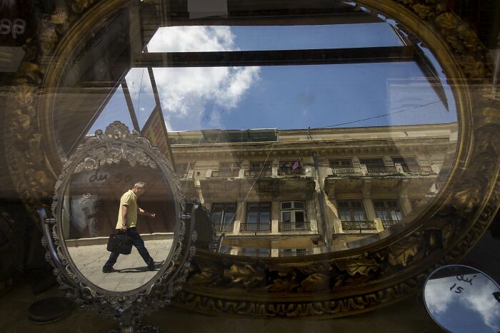 Man walking past an ornate mirror on a busy street, creating a humorous scene with a building reflection in the background.