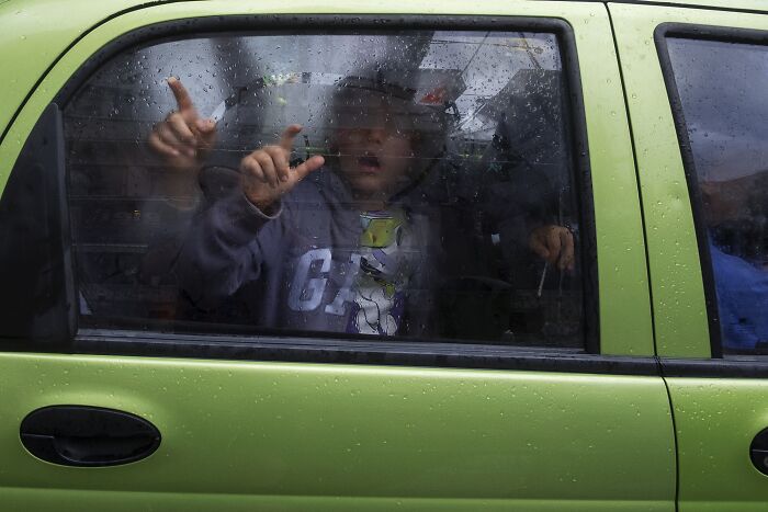 Child making funny faces through a rainy car window, capturing a perfectly timed street photo moment.