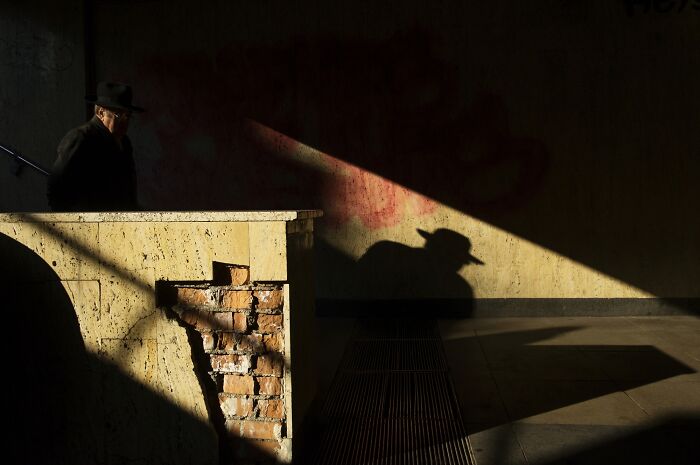 Shadow of a man with a hat on a building wall at sunset, creating a comedic effect in a street photo by Cosmin Gârleșteanu.