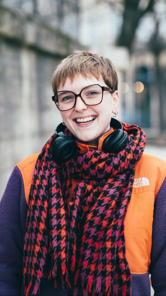 Smiling person in glasses, colorful scarf, and headphones; vibrant street portrait by photographer.