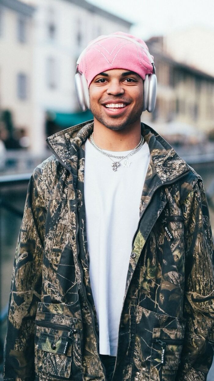 Street portrait of a smiling person in a pink hat and camo jacket with headphones, captured by a photographer.