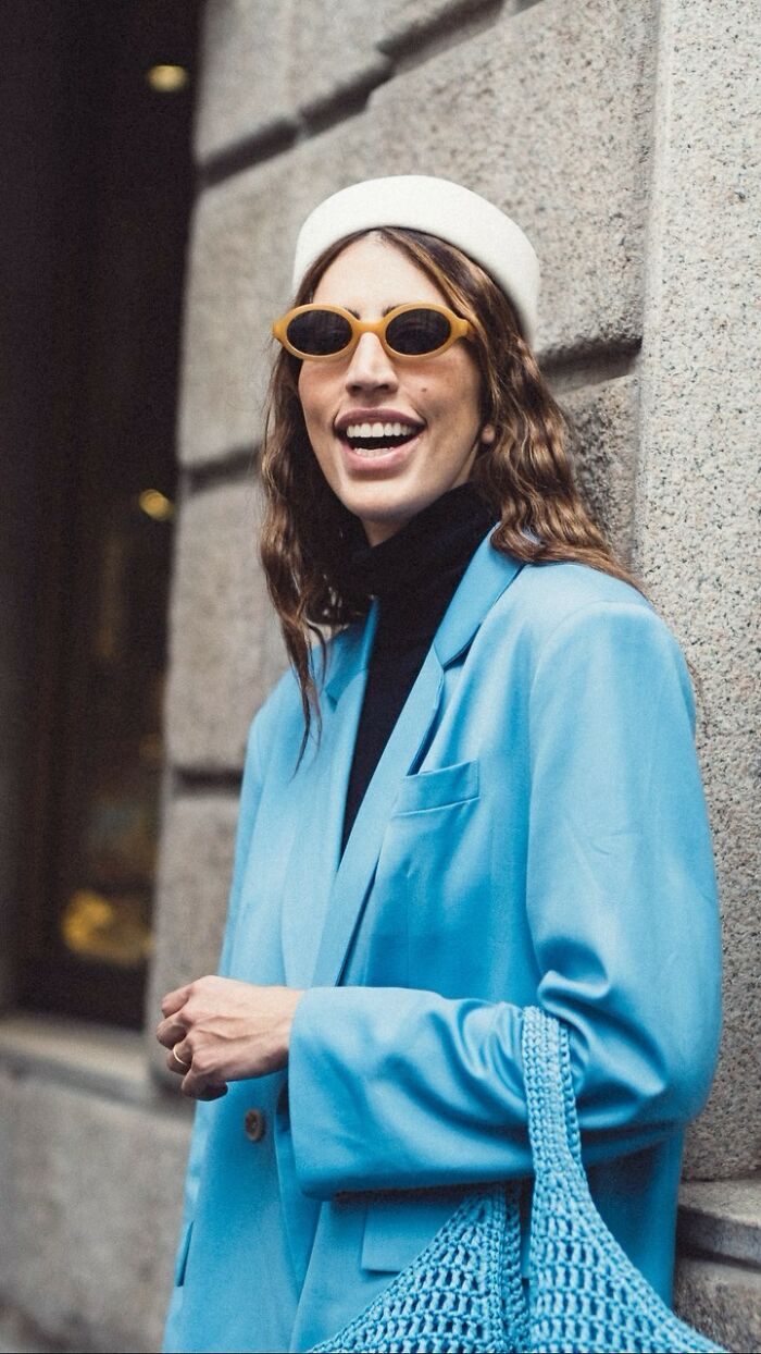 A street portrait of a smiling woman in a blue blazer, wearing sunglasses and a white beret, leaning against a stone wall.