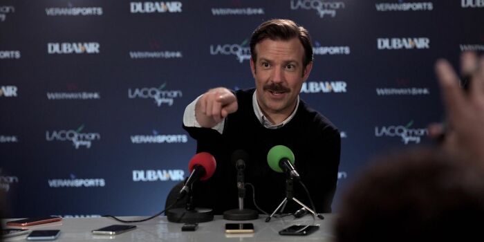 Actor at a press event, pointing towards the audience, with two microphones in front of him.