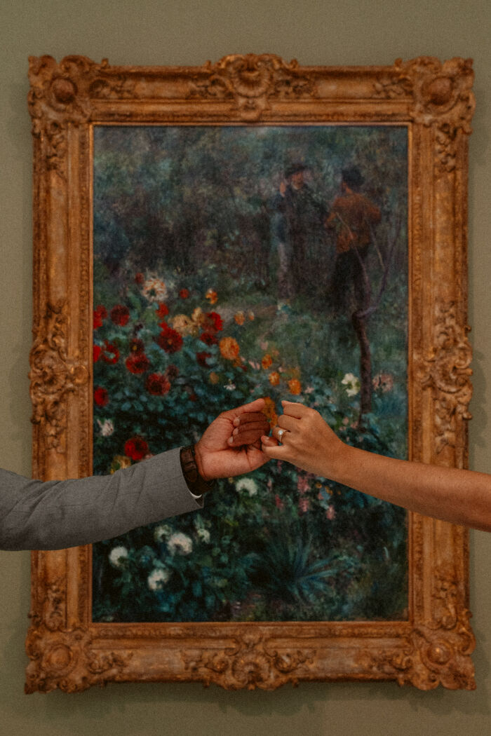 Engagement photo featuring hands with a ring, set against an ornate framed painting background.