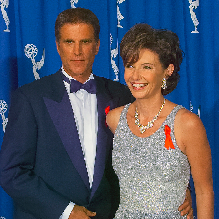 Ted Danson and Mary Steenburgen poses at an event, man in a tuxedo and woman in a sparkly gown, both with red ribbons.