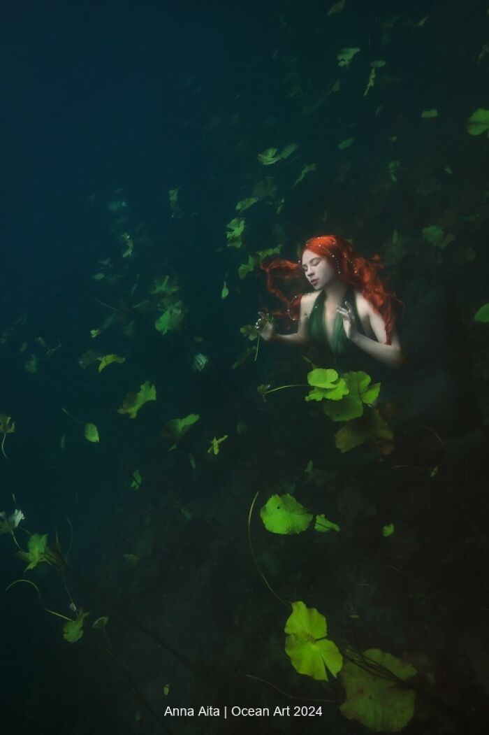 Underwater art photo of a figure with red hair surrounded by green leaves, conveying a serene, mystical scene.