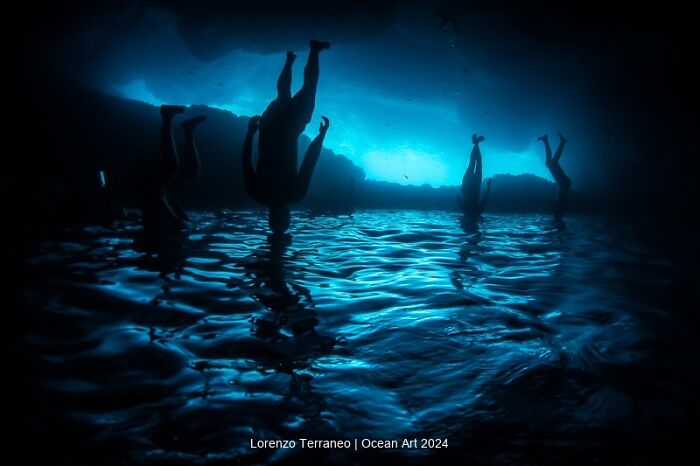 Underwater photo of silhouetted divers upside down in a blue-lit cave, Ocean Art Photo Contest 2024 winner.