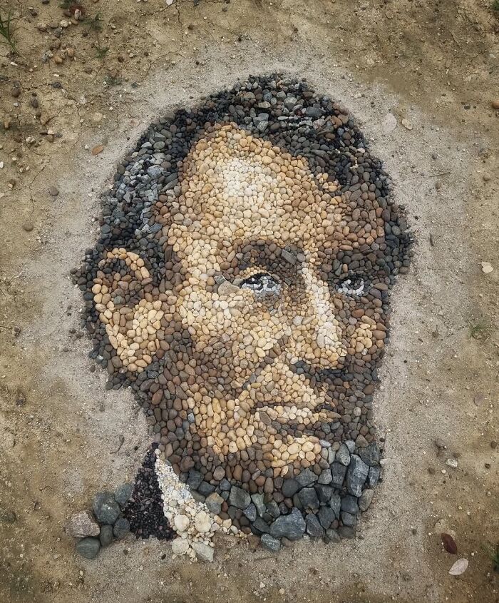 Intricate stone and pebble mosaic portrait on sandy ground, showcasing British artist's temporary art style.