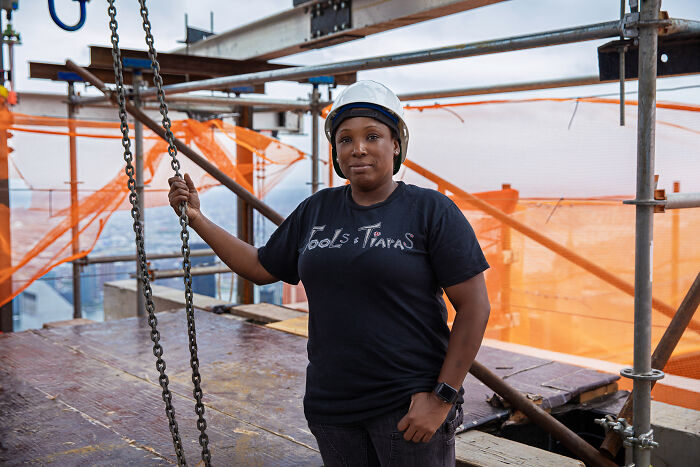 A woman in construction gear stands confidently on a building site, challenging society's norms.