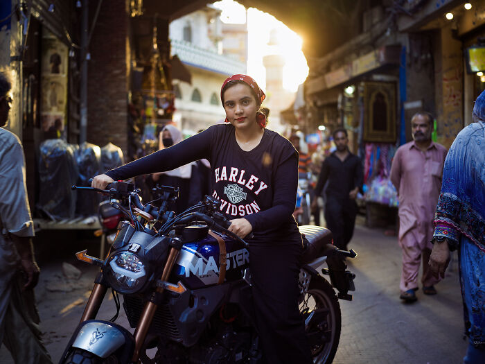 A woman on a motorcycle, confidently posing in a busy market, defying traditional societal norms.