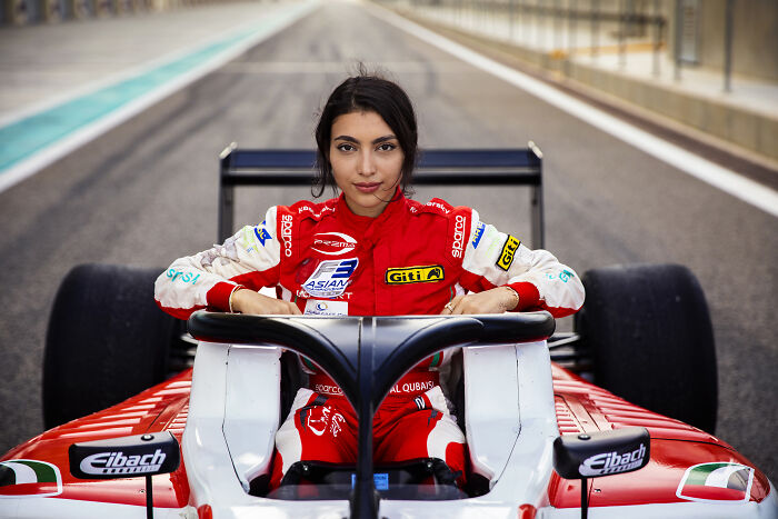 Woman race car driver in red suit, challenging societal norms, seated in a red car on a racetrack.