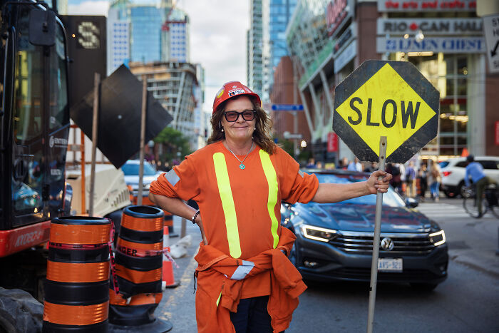 Woman in construction gear holding a "SLOW" sign in a city, showcasing defiance against societal norms.