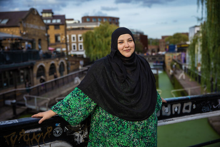 Woman defying societal norms, standing on a bridge in a green outfit, with a canal and buildings in the background.