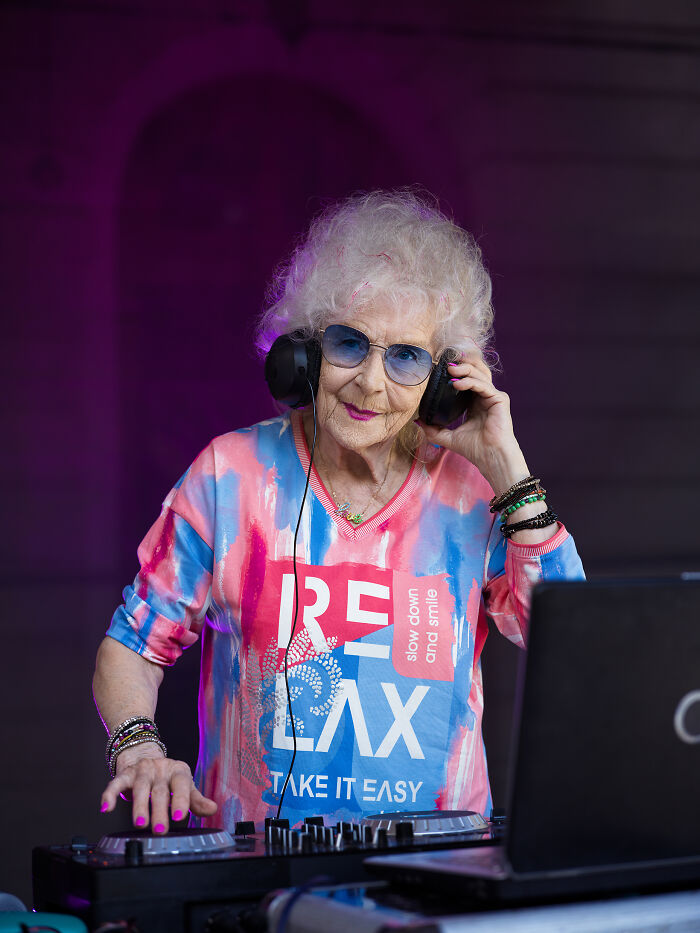 Elderly woman DJ defying societal norms, wearing headphones and a colorful top in a vibrant setting.