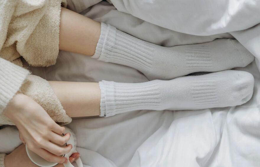 Person wearing cozy socks and holding a mug, relaxing in bed, suggesting nighttime showering benefits.