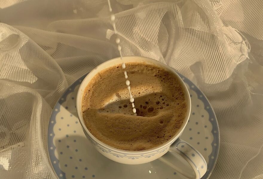 Milk pouring into a coffee cup on a saucer, surrounded by soft white fabric.