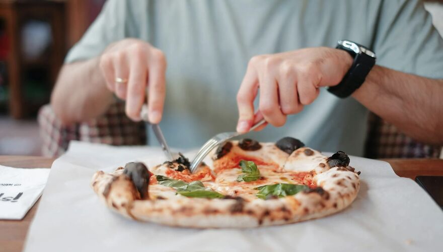 Man enjoying pizza at a table, wearing a smartwatch, with a casual setting in the background.