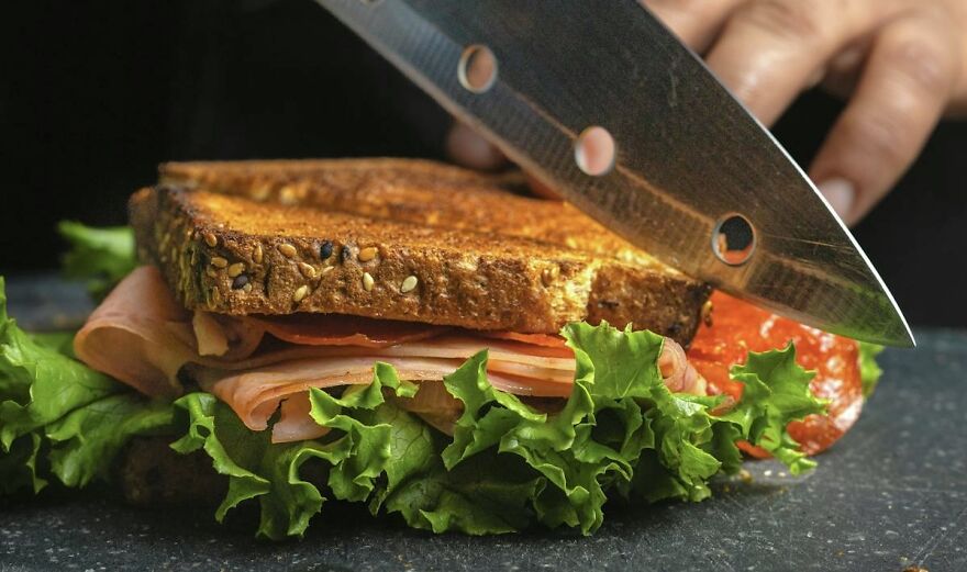 A sandwich with lettuce and tomatoes being sliced with a knife on a cutting board, highlighting the best way to do things.