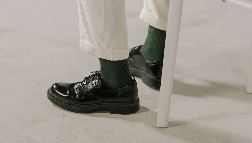 Person wearing white pants and black shoes with green socks, seated, feet view.