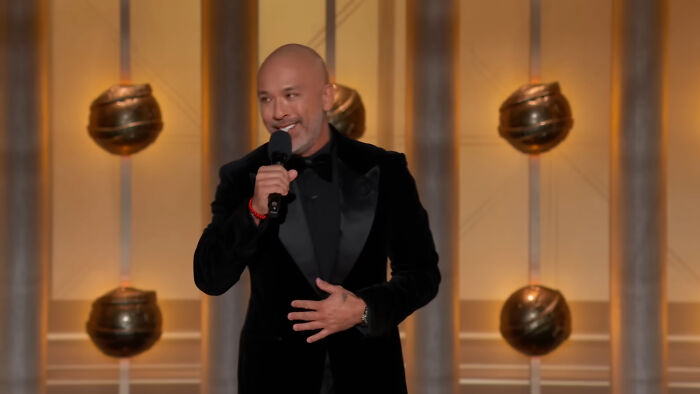 Man in a black suit holding a microphone on stage during a scandalous awards show moment.